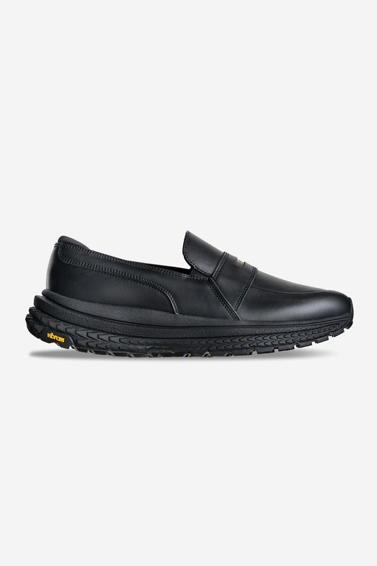 The Liaizon Vibram - Black is a black leather slip-on loafer with sleek design, visible stitching, a thick textured rubber sole, and a small yellow Vibram logo near the heel.