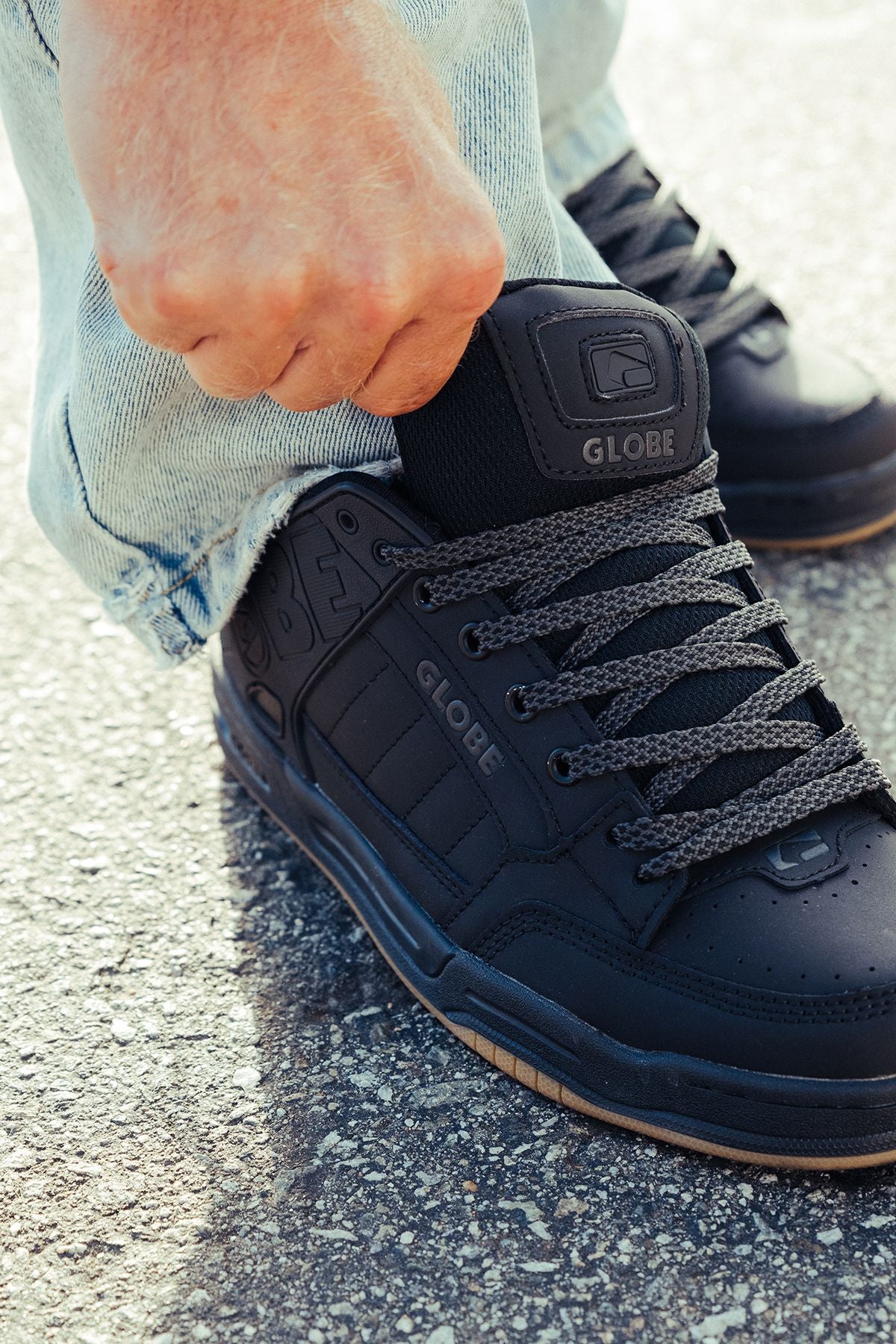 A person in light blue jeans is pulling up the tongue of a Tilt - Dark Shadow/Phantom skate shoe with gray laces while standing on textured pavement.