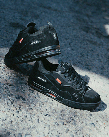 A pair of black athletic sneakers with red and white accents are displayed on a textured, sunlit pavement. One shoe is upright while the other leans on its side, showcasing the side and sole details.