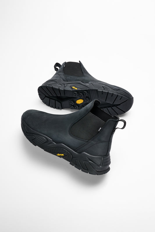 The DOVER-2000 - Black/Black is a pair of black Chelsea boots made from crazy horse leather, with thick rugged Vibram soles featuring yellow branding and pull tabs at the back, shown on a white background.