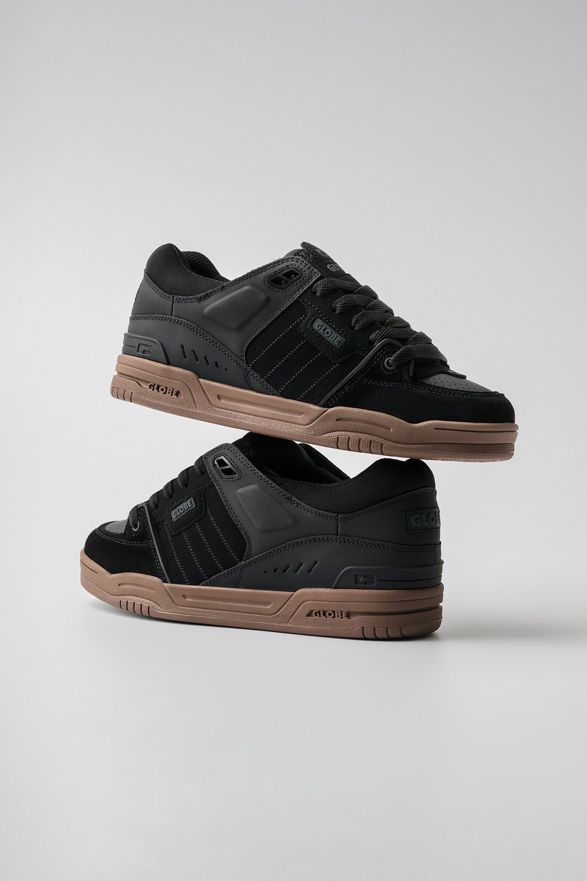 The Fusion - Black/Tobacco Gum: a pair of low-top sneakers in black and dark grey with brown soles, one shoe atop the other, shown on a plain light gray background.