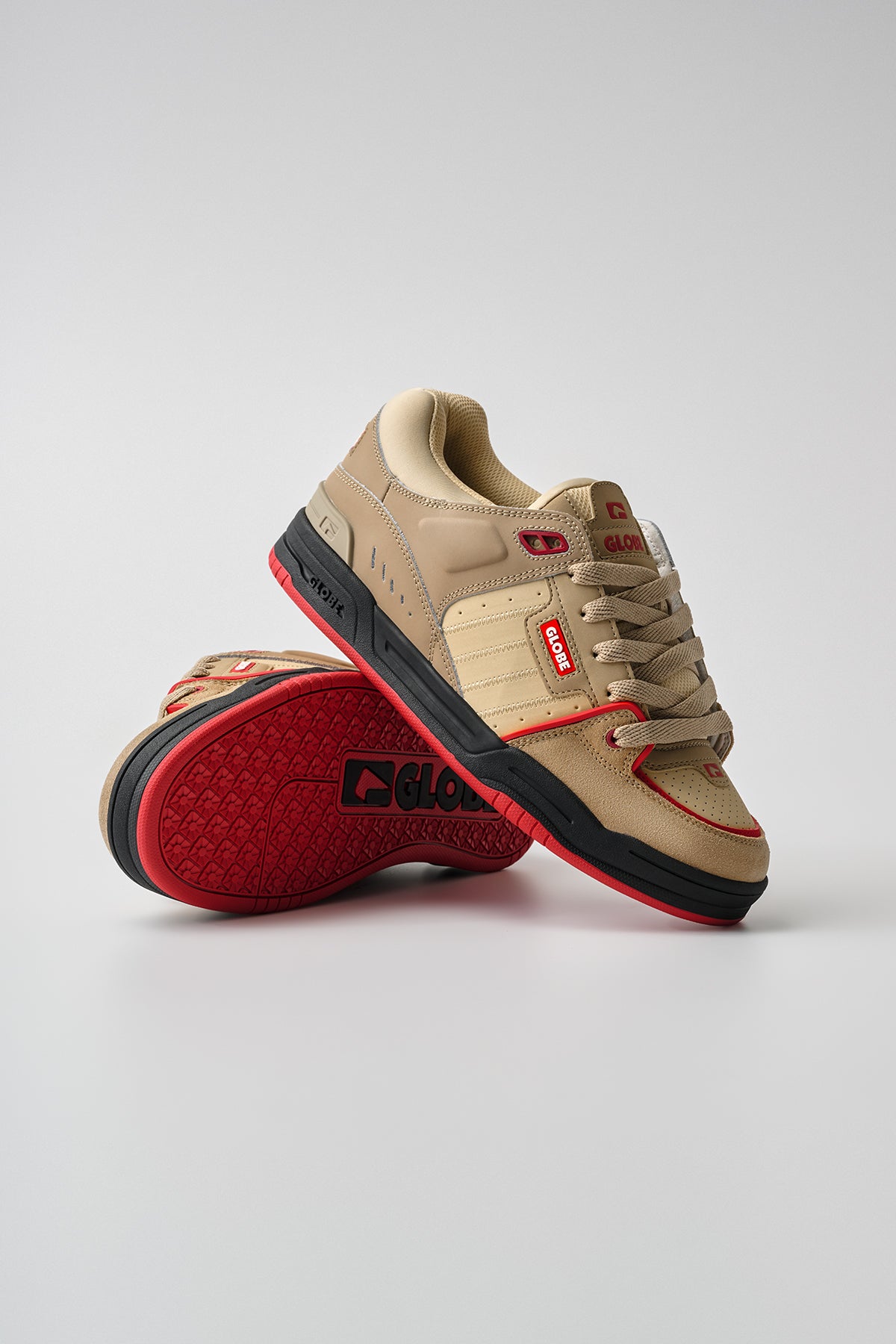 A pair of Globe Fusion - Khaki/Red skate shoes with red accents and black soles; one stands upright, the other lies on its side, revealing the red tread with the Globe logo. Set against a plain light background.
