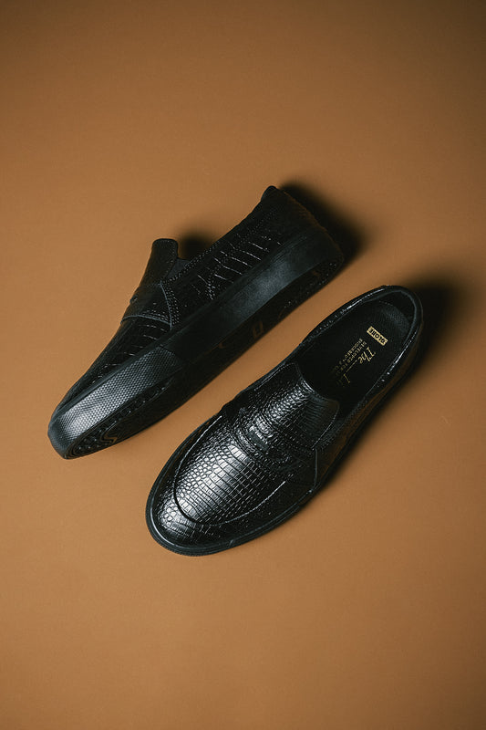 Two black crocodile texture Liaizon loafers on a warm brown backdrop, one showing the insole label