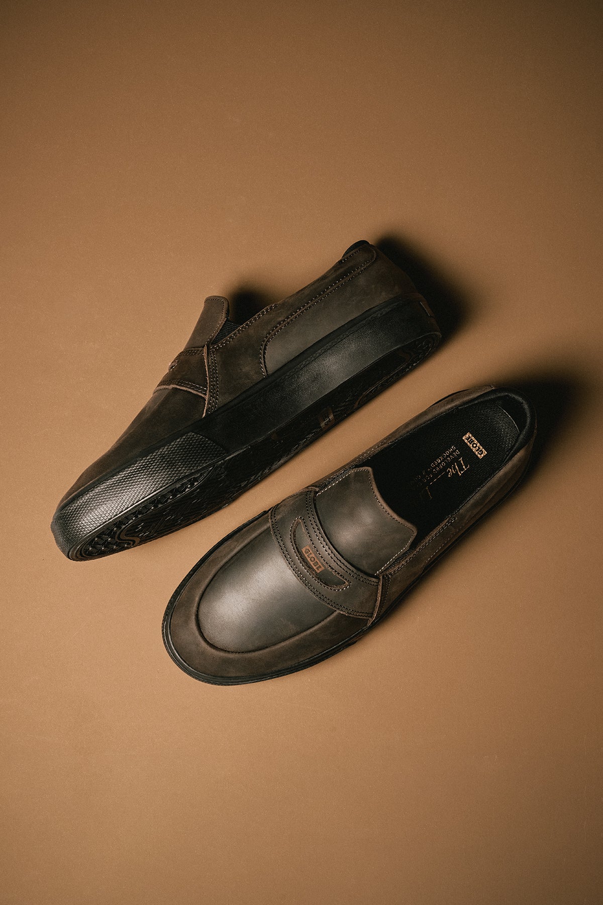 Diagonal shot of brown Liaizon loafers on a tan backdrop highlighting the classic slip-on silhouette and stitching