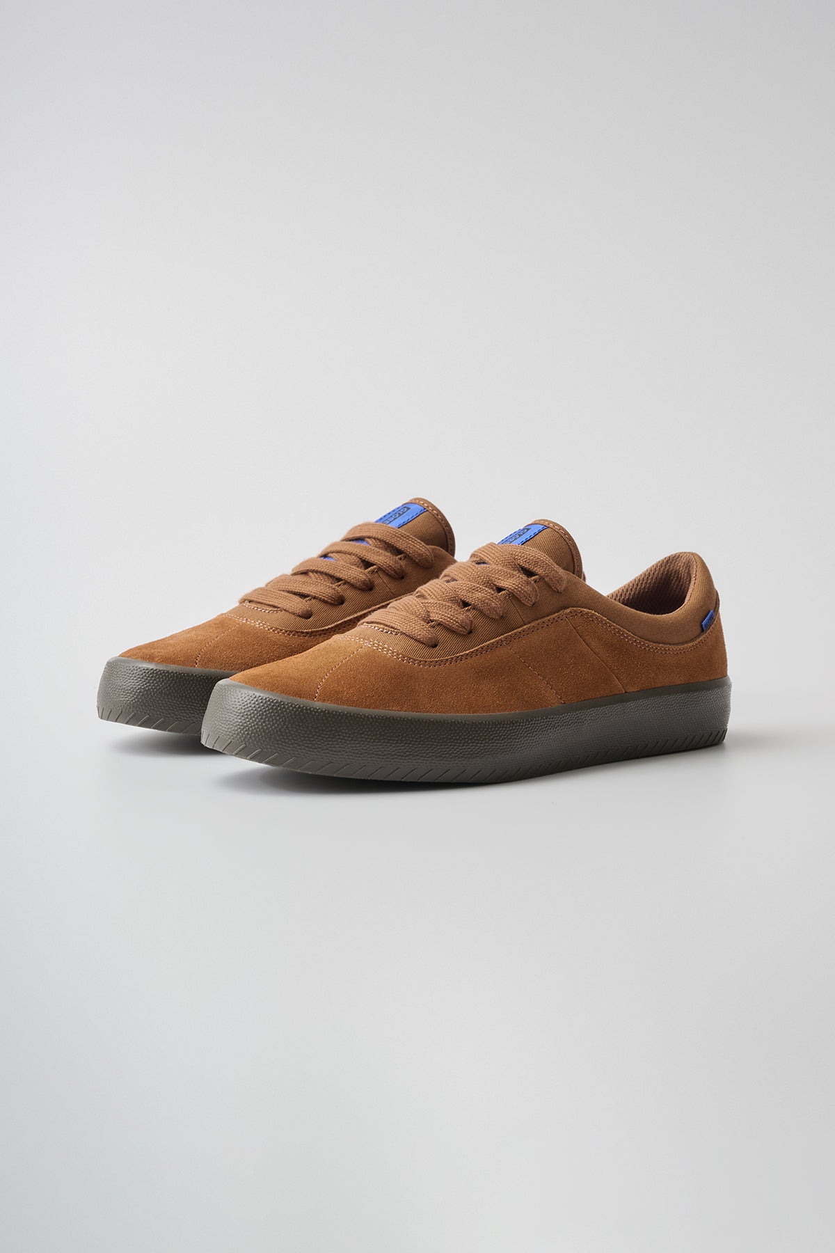 The Melbek - Caramel sneakers feature brown suede uppers, brown laces, thick dark brown rubber soles, and a small blue tag on the tongue and heel, displayed on a light gray background.