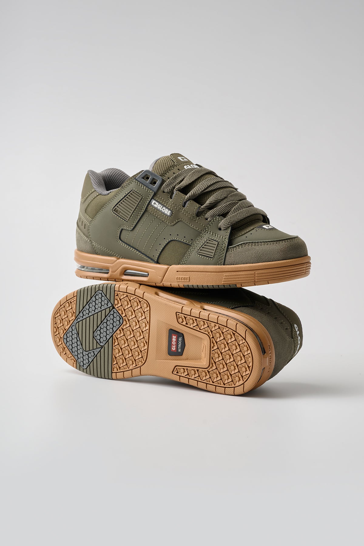 The Sabre - Moss/Gum features olive green low-top sneakers with gum soles. One stands upright, the other on its side showing tread and branding. Laced with white logo details.