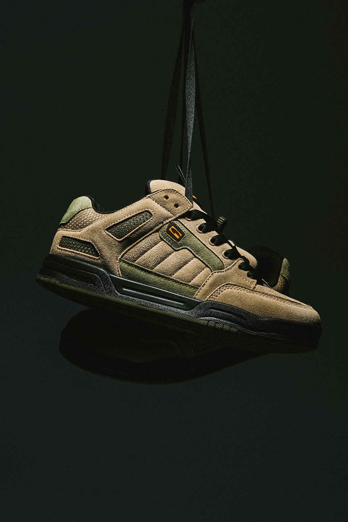 Tilt Prime skate shoe in olive and orange tones suspended by a strap against a dark background