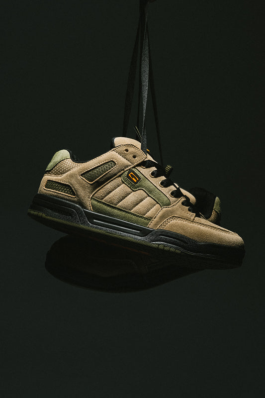 Tilt Prime skate shoe in olive and orange tones suspended by a strap against a dark background