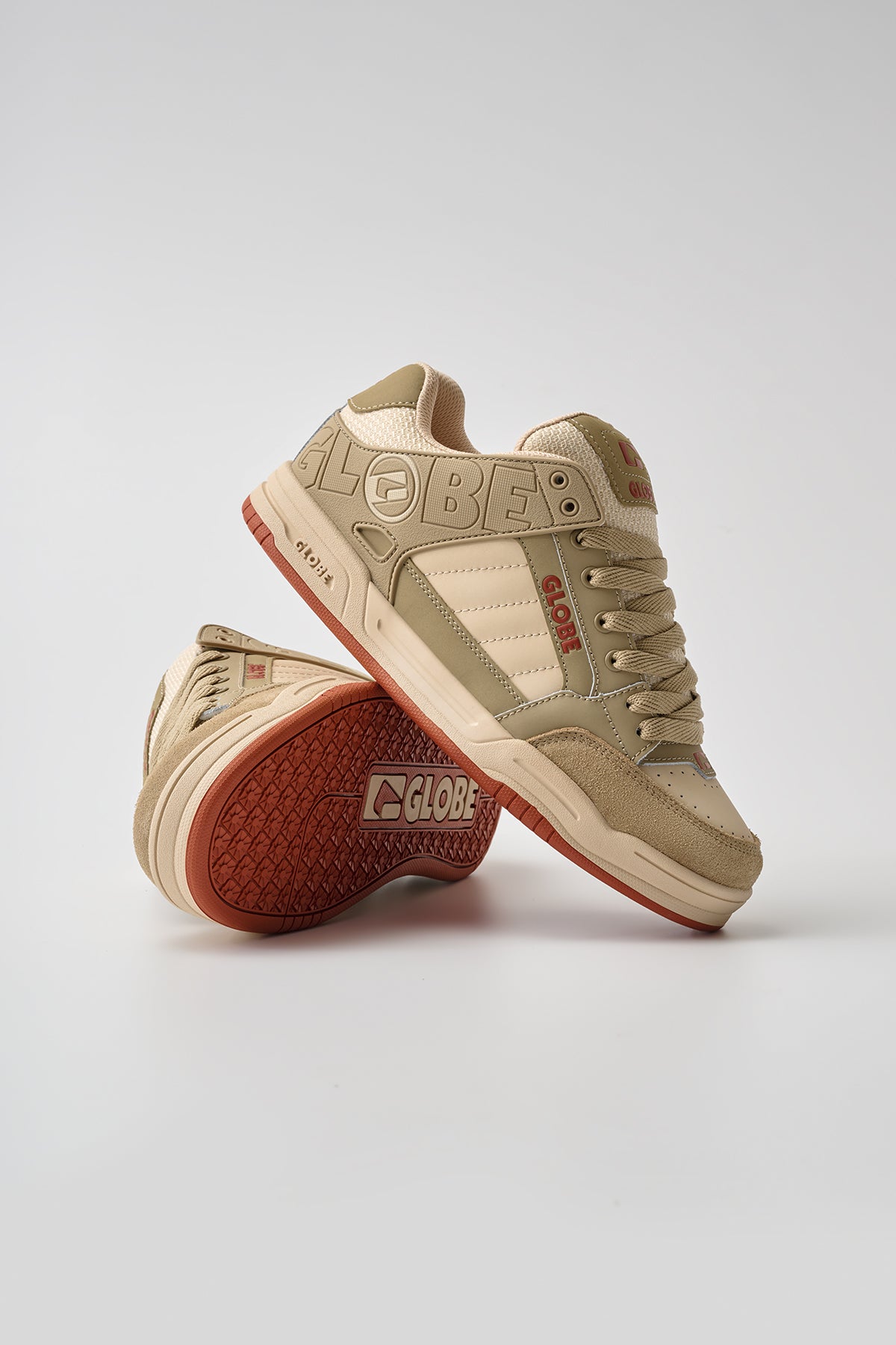 A pair of Globe Tilt skate shoes in Sand/Red, featuring beige uppers and red-brown soles with the Globe logo. One shoe stands upright while the other leans to display its sole against a plain light gray background.