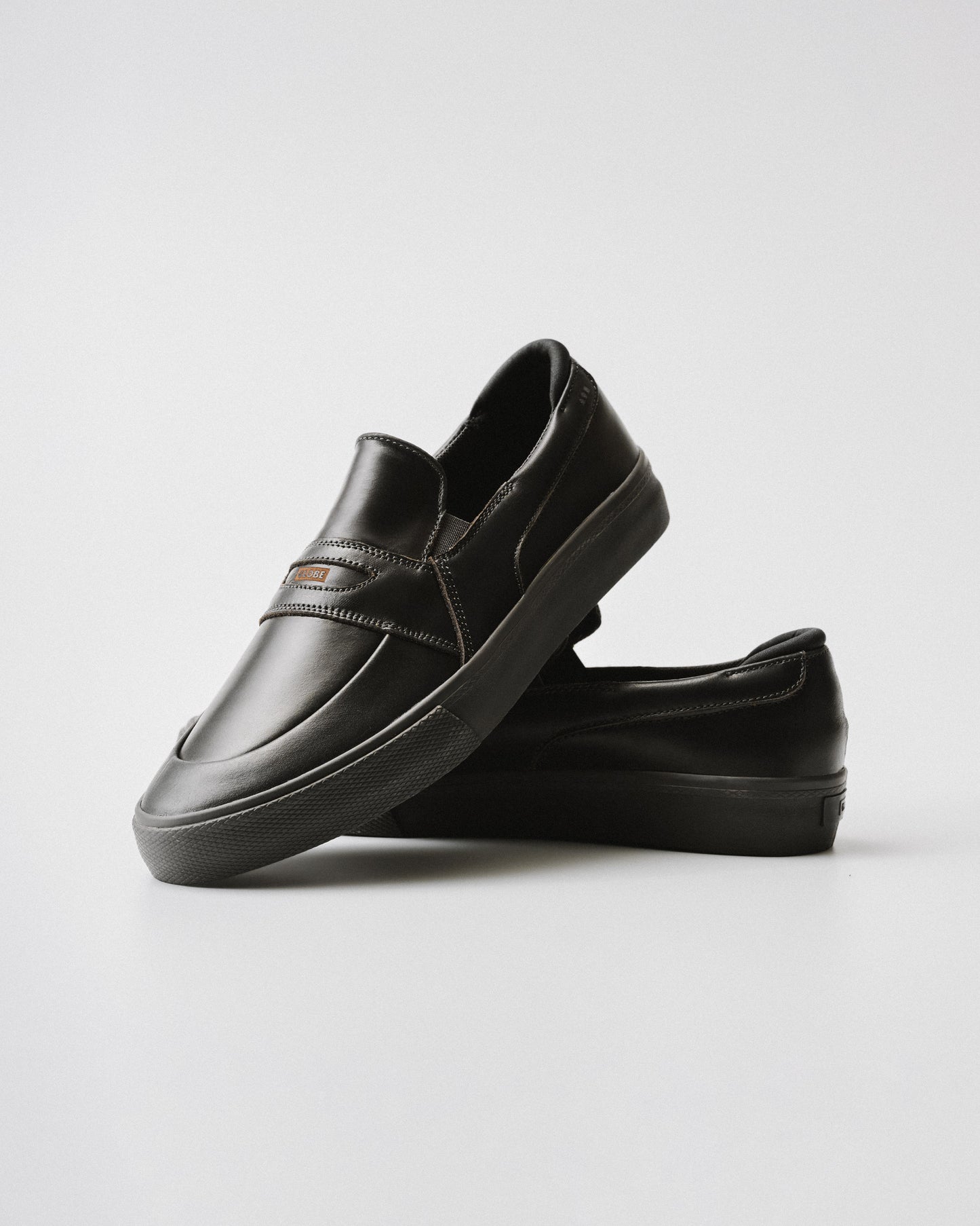 The Liaizon - Black/Maalouf skate shoes, crafted from premium pull-up leather, are shown in black—one upright and one on its side—against a plain white background.