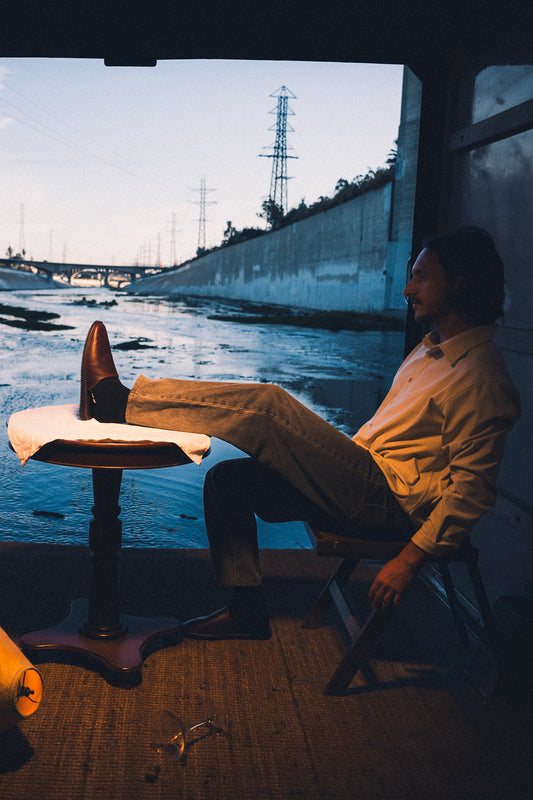 A man wearing Mule - Oxblood/Gillette leather mules with Vibram soles sits on a wooden chair by an urban river, one foot on a white-draped table, gazing out as dusk falls alongside power lines and a concrete embankment.