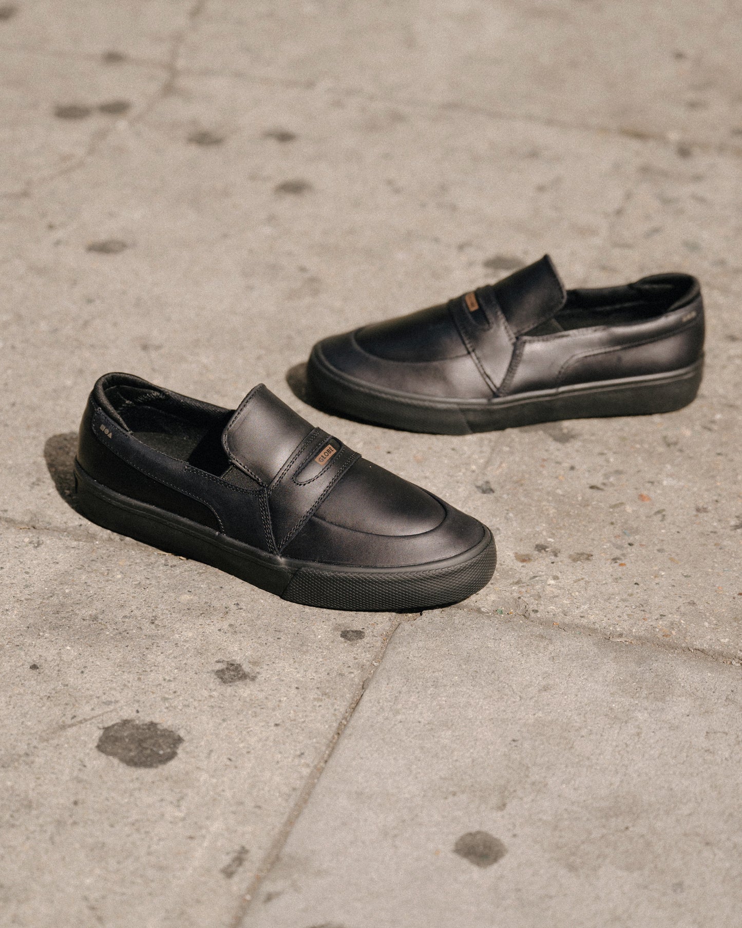 A pair of Liaizon - Black/Maalouf skate shoes with rubber soles rests on a concrete sidewalk, one upright and the other on its side.
