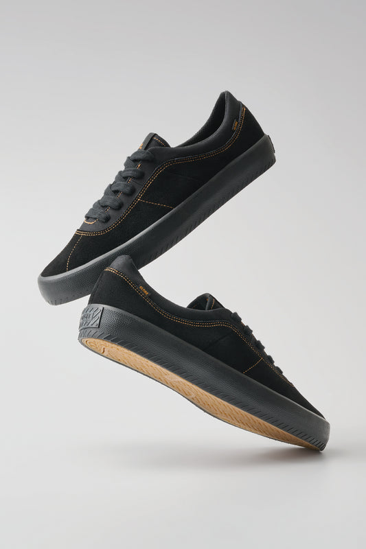 The Melbek - Black/Montano skate shoes, featuring a suede upper, orange stitching, and textured soles, float in mid-air against a light gray background.