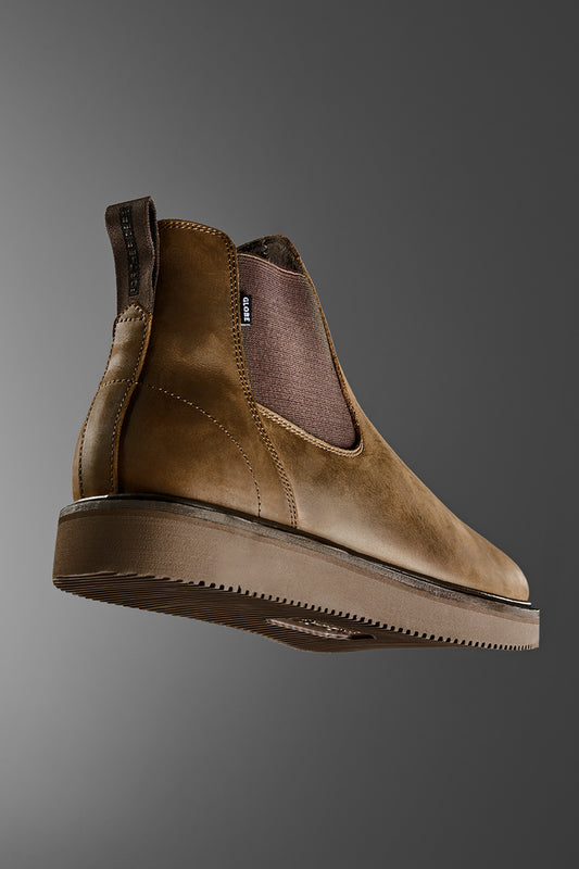 The Dover II Vibram - Brown Crazyhorse Chelsea Boot features brown leather, an elastic side panel, and pull tab. Shown on a gray background from a low angle, it showcases its sleek design and durable Vibram outsole.