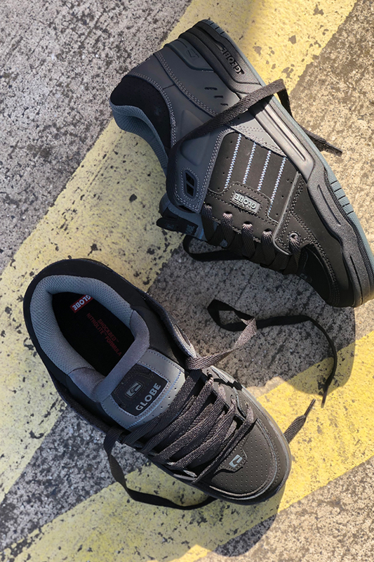 Two Globe Fusion - Black/Night leather skate shoes with gray accents and S-Trac soles rest on a weathered concrete surface marked by yellow lines. One stands upright and the other lies on its side, both featuring untied laces.
