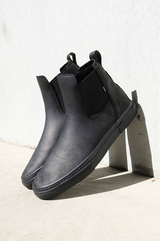 The Dover II - Black Crazyhorse boots feature black leather, elastic side panels, and pull tabs. Styled like a Chelsea boot yet versatile for skateboarding, they're shown upright against a white wall on a concrete surface.