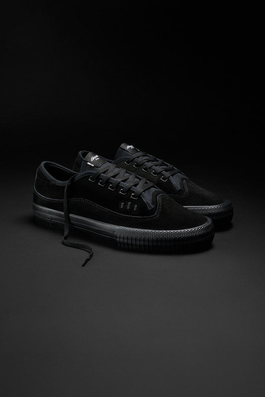 The Gillette - Black/Suede/Black skate shoes feature black suede, black laces, a Lux PU insole, and subtle stitching. One shoe with an untied lace rests on a dark background.