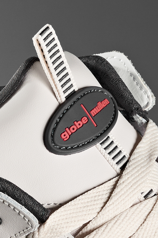 Close-up of the Globe Tilt Prime - Cement / Mullen skate shoe, inspired by Rodney Mullen. Features a black oval patch with red text, white laces and upper, plus black and white striped pull tabs—ideal for street skating.