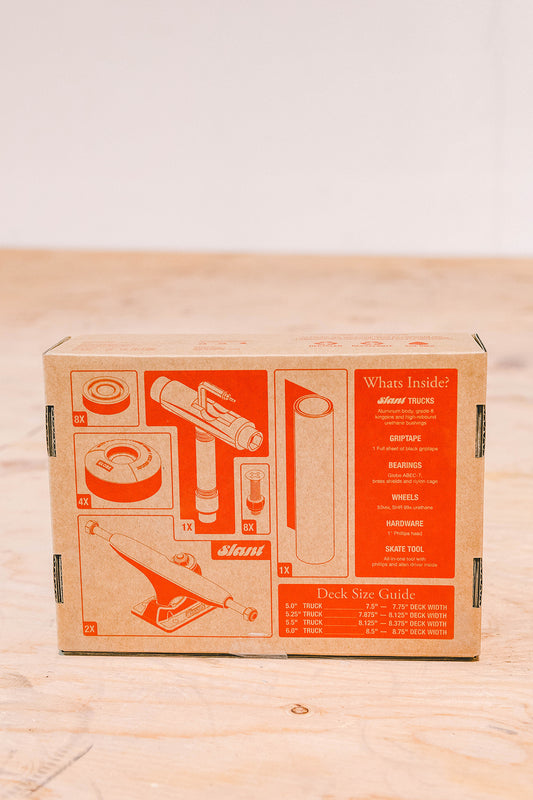 A cardboard box labeled "Everything But The Deck Kit Accessories Hg 5.0" - Raw Finish," featuring orange graphics, contains Slant trucks, grip tape, wheels, bearings, hardware, and a skate tool on a wooden surface.