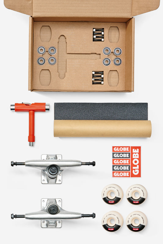 The Everything But The Deck Kit Accessories Hg 5.0" - Raw Finish includes Slant trucks, cream wheels, eight bearings, grip tape, a T-tool, hardware, and red Globe stickers—neatly arranged below an open cardboard box.