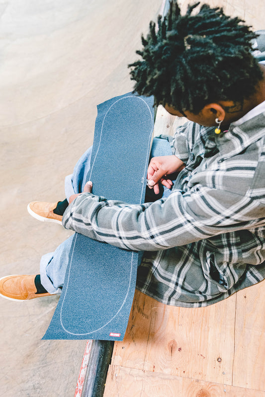 A person with short dreadlocks in a plaid shirt and tan shoes sits on a skate ramp, applying grip tape to a skateboard deck, surrounded by parts from the Everything But The Deck Kit Accessories Hg 5.0" - Raw Finish.
