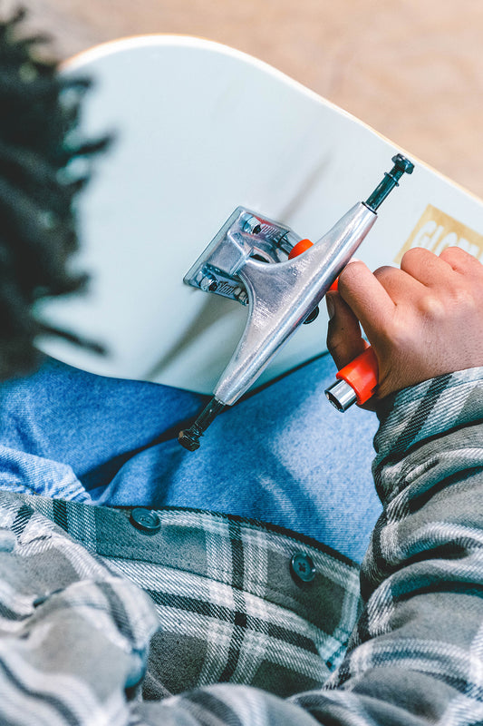 A person wearing a plaid shirt and jeans uses an orange tool to attach Everything But The Deck Kit Accessories Hg 5.0" - Raw Finish trucks to a white skateboard deck, assembling essential skateboard parts.