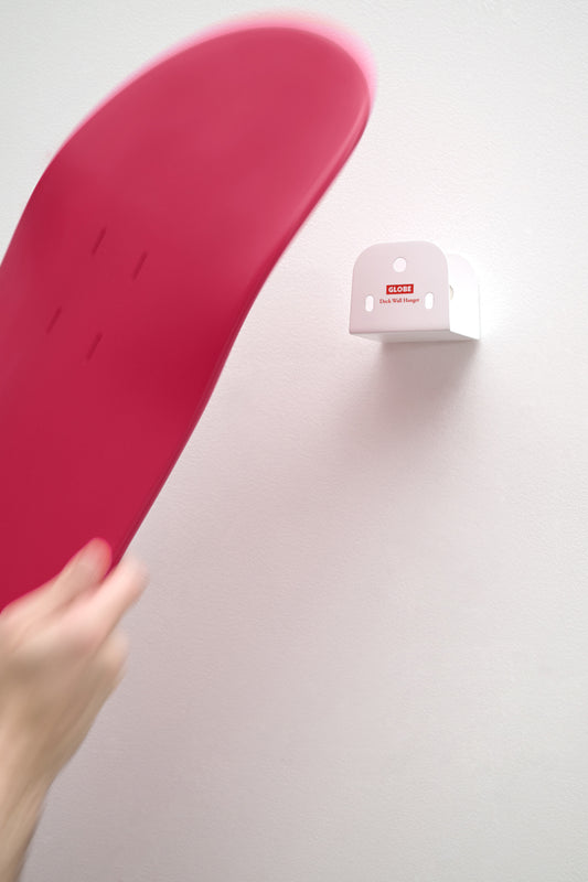 A person holds a red skateboard deck near the white "Skateboard Deck Wall Hanger Accessories Hg 1sz" mount, featuring a red CLOSE label. The plain white background highlights the hanger’s simple, modern design.