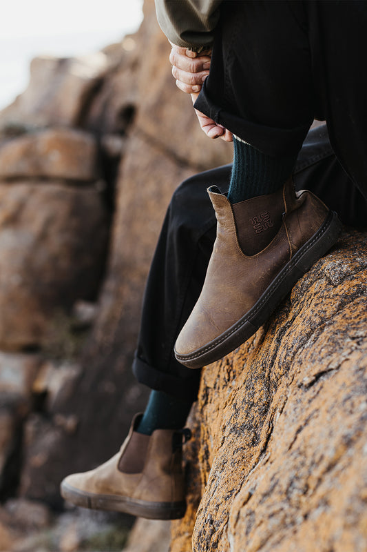 Wearing Dover II - Dark Brown/Wasted Talent Chelsea boots, black pants, and dark green socks, a person sits on rocky terrain with one leg bent and hands on knee; rugged rock surface features in the background.