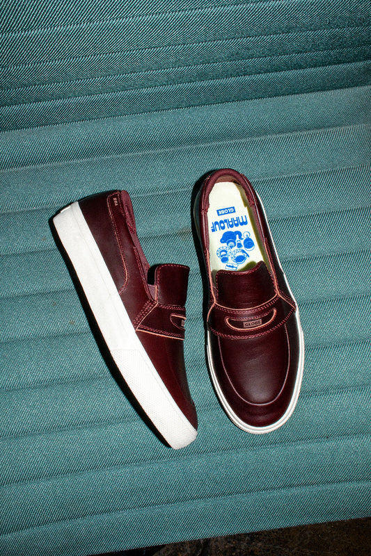 A pair of Liaizon - Oxblood/Maalouf skate slip-ons with white soles sits on a textured teal surface. One shoe is upright, the other on its side, showing cartoon-graphic insoles and branding.